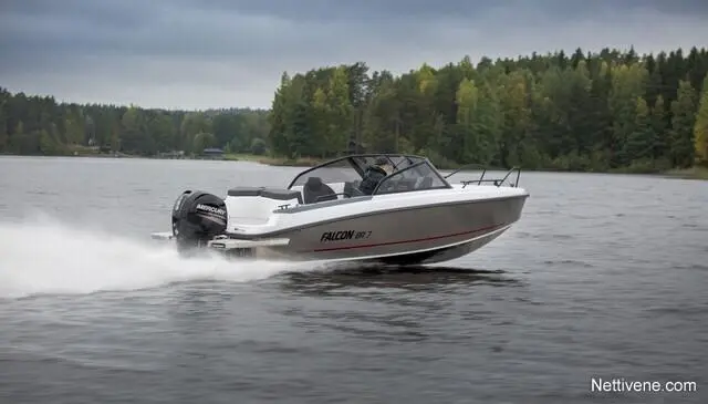 A white Falcon 590 boat speeds across a calm lake, leaving a wake, surrounded by green trees under a cloudy sky.